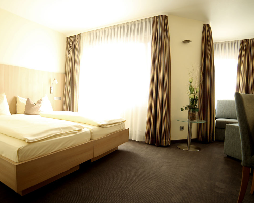 Ein modern eingerichtetes Hotelzimmer mit Doppelbett, Sessel, Tisch mit Blumen und großen Fenstern mit Vorhängen.